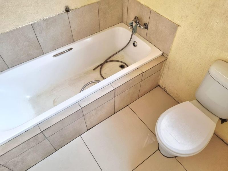 A functional bathroom featuring a built-in bathtub with shower mixer and a standard toilet, finished with neutral floor and wall tiling.