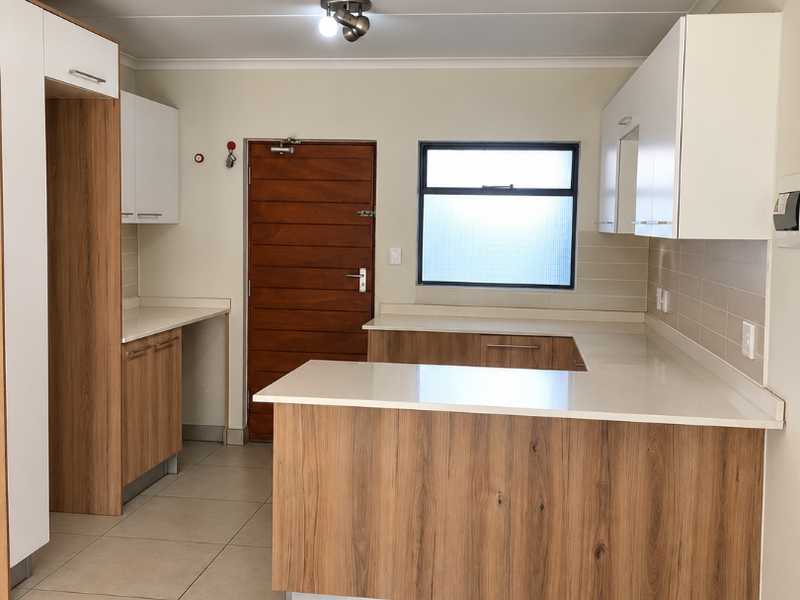 A modern, clean kitchen featuring light wood-grain cabinetry, white stone countertops, tiled flooring, and a neutral backsplash, presenting a bright and functional workspace.