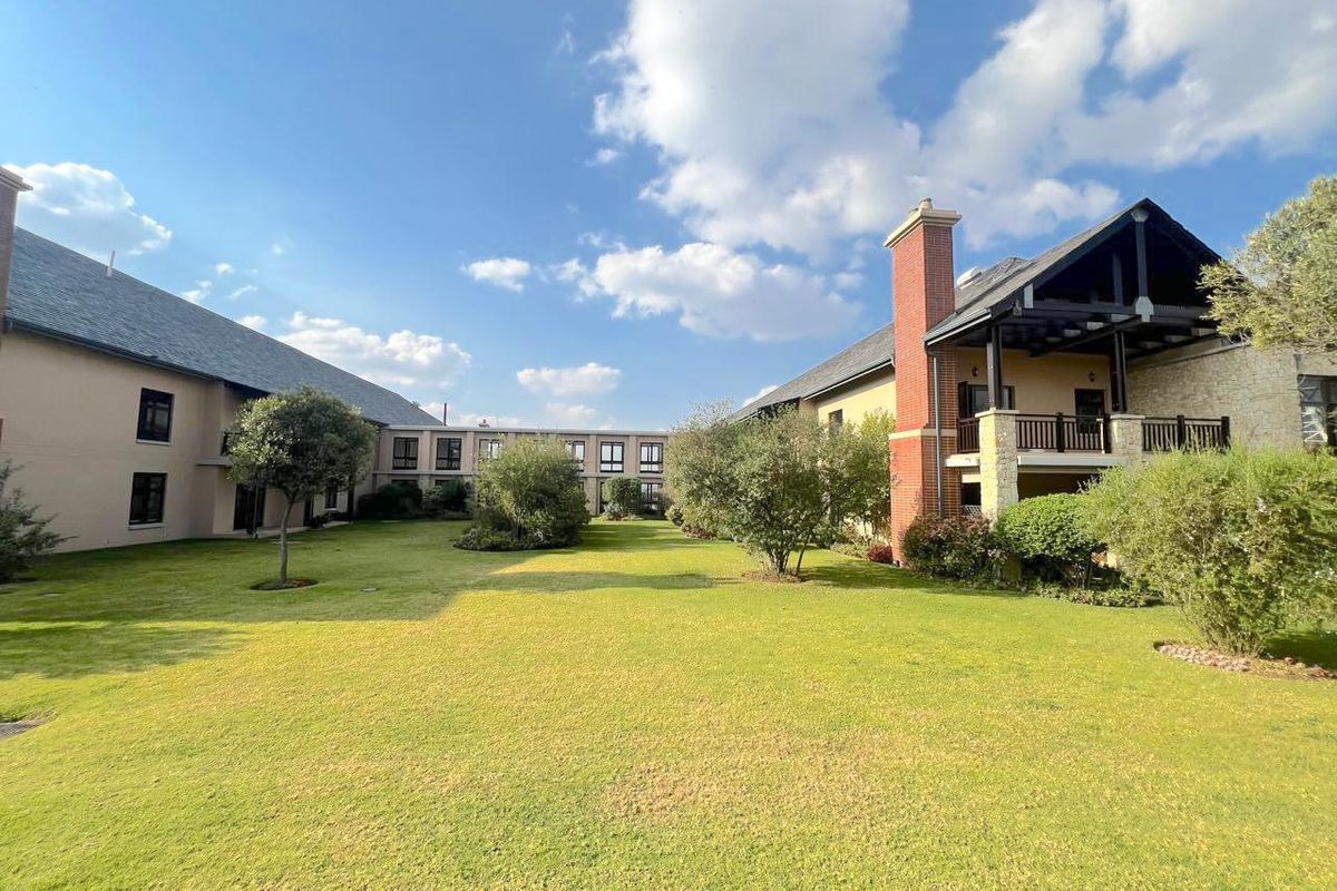 A well-maintained communal garden area featuring manicured lawns, mature trees, and views of the retirement village building architecture with balconies and brick chimneys.