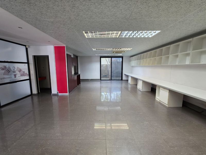 Spacious open-plan office area featuring built-in desk workstations, extensive wall shelving, tiled flooring, and a drop ceiling with integrated lighting.