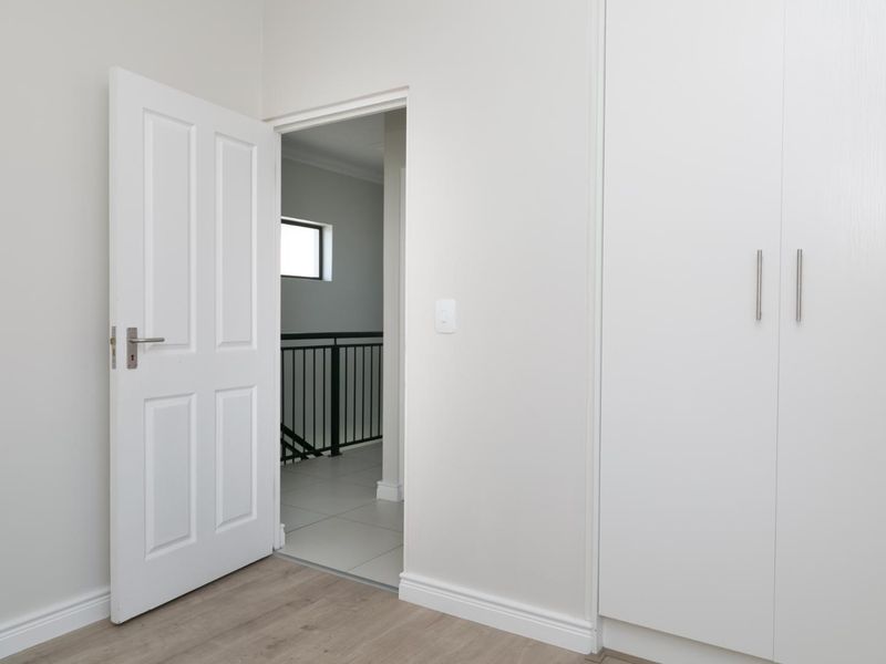 A clean, modern bedroom featuring neutral walls, laminate flooring, and built-in cupboards, with a view into the adjacent hallway.