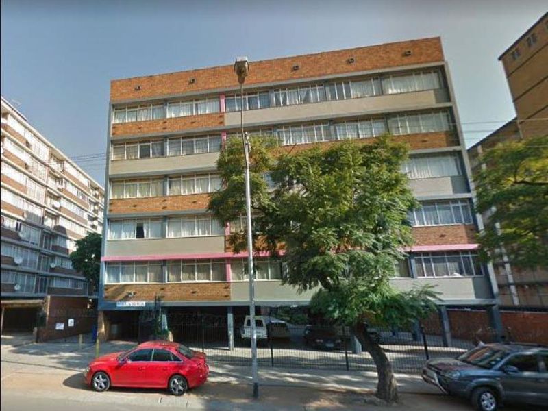 Front elevation of a multi-story apartment building featuring a brick and concrete facade with ground-level parking and security fencing.