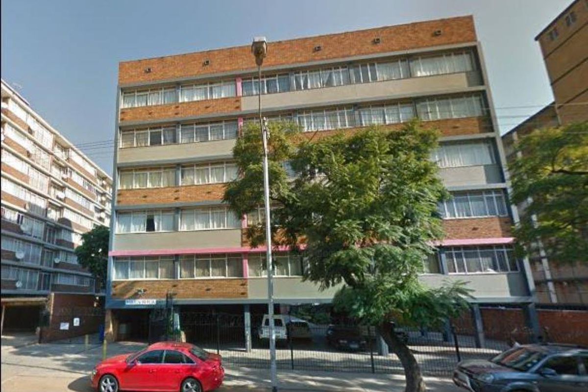 Front elevation of a multi-story apartment building featuring a brick and concrete facade with ground-level parking and security fencing.