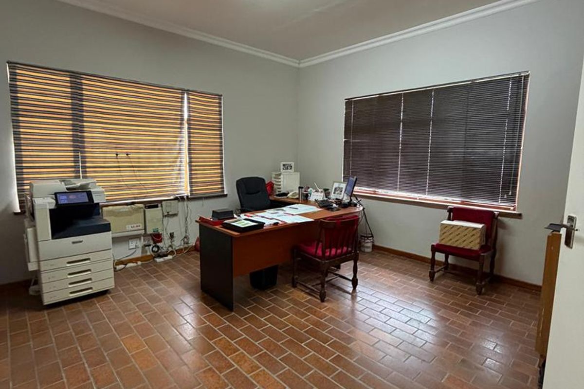 A functional office space featuring terracotta tile flooring, large windows with venetian blinds, and overhead fluorescent lighting.