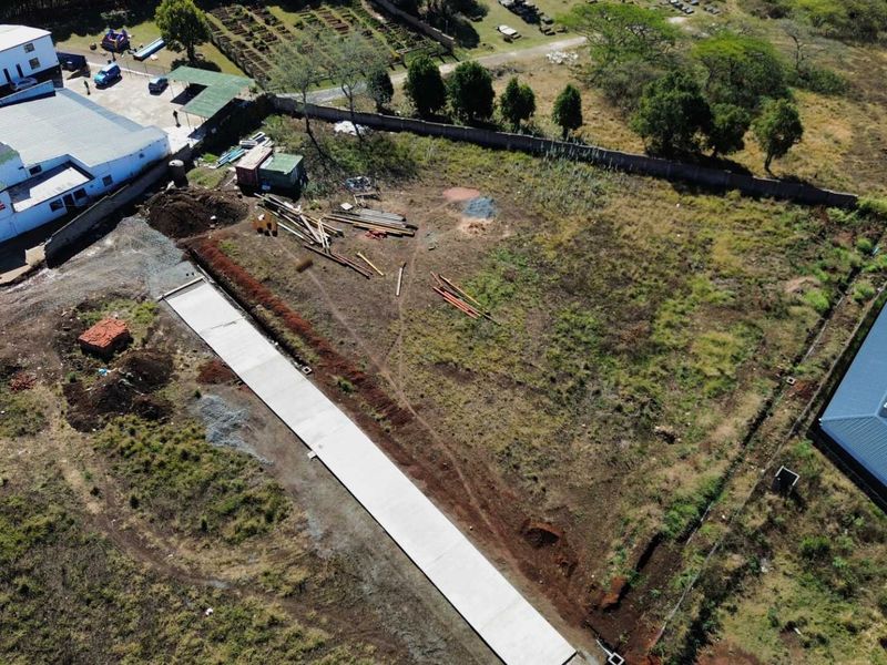 Aerial view of a vacant commercial land plot featuring a newly laid concrete driveway and perimeter walling, situated adjacent to existing commercial structures.
