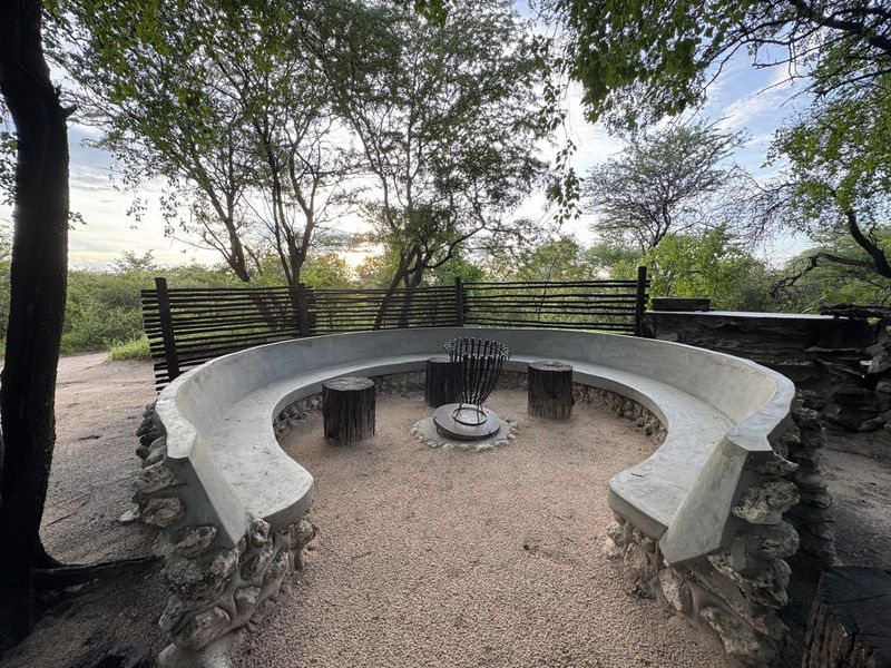 An outdoor boma area featuring a built-in circular concrete bench, stone masonry detailing, and a central fire pit set within a natural garden.
