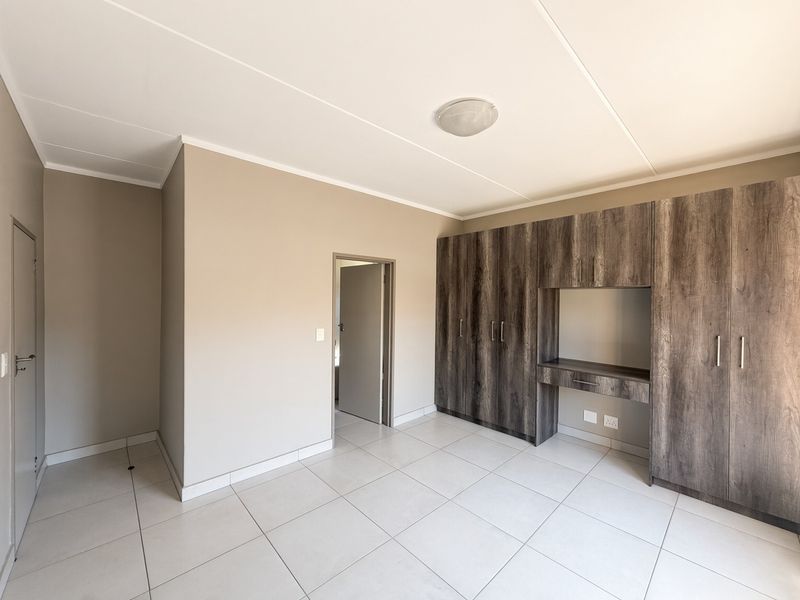 A clean, empty bedroom featuring neutral wall tones, modern tiled flooring, and extensive built-in wooden cabinetry with an integrated desk area.