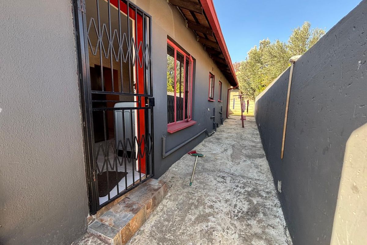 A narrow side exterior walkway featuring concrete paving, security gate, and dark grey painted exterior walls with red window frames.