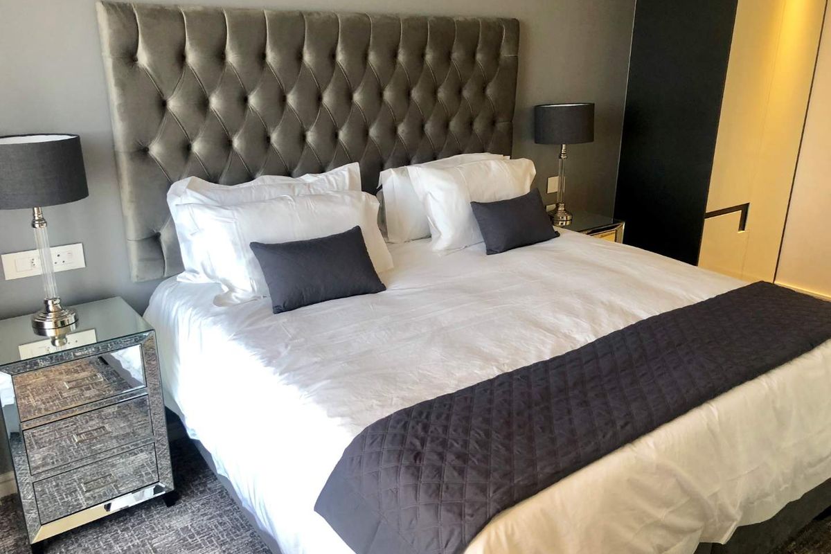 A well-appointed bedroom featuring a large tufted headboard, neutral grey walls, and integrated storage solutions in a modern, clean aesthetic.