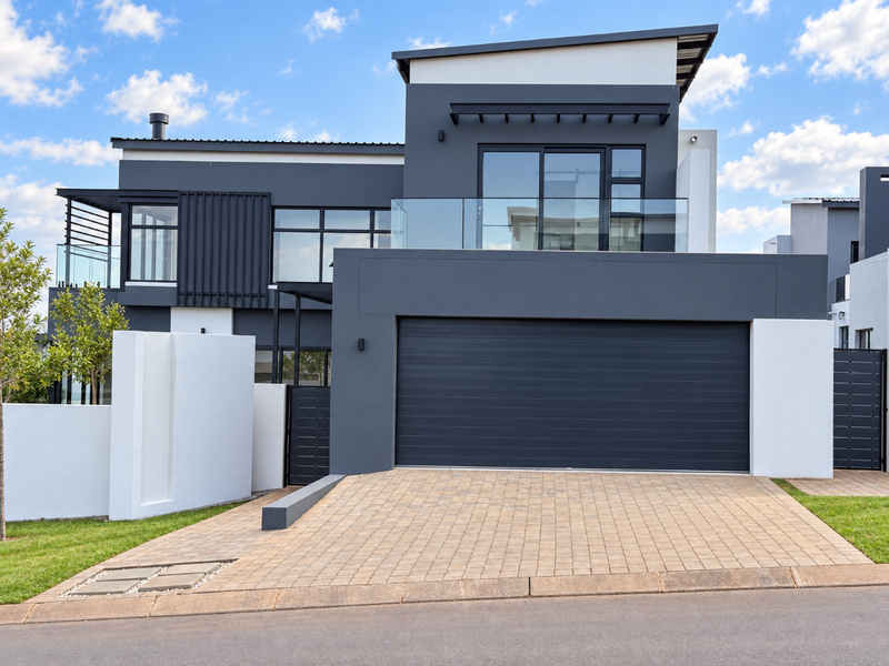 Modern double-story home featuring a sleek dark grey and white facade, paved driveway, and a spacious double garage with a glass-railed balcony above.