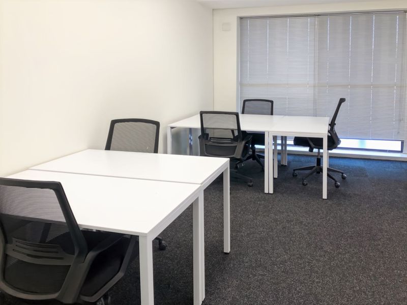 A clean, functional office space featuring neutral walls, dark carpet flooring, and large windows fitted with adjustable venetian blinds.