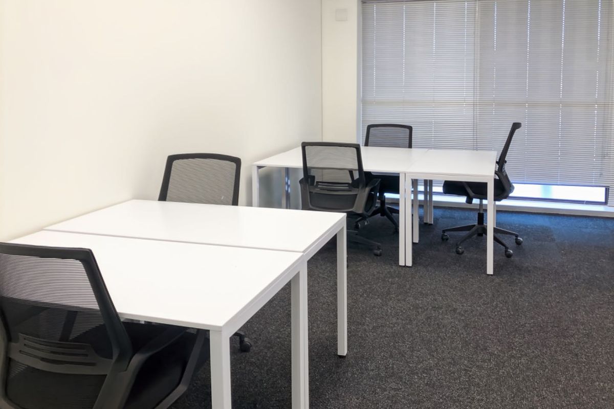 A clean, functional office space featuring neutral walls, dark carpet flooring, and large windows fitted with adjustable venetian blinds.