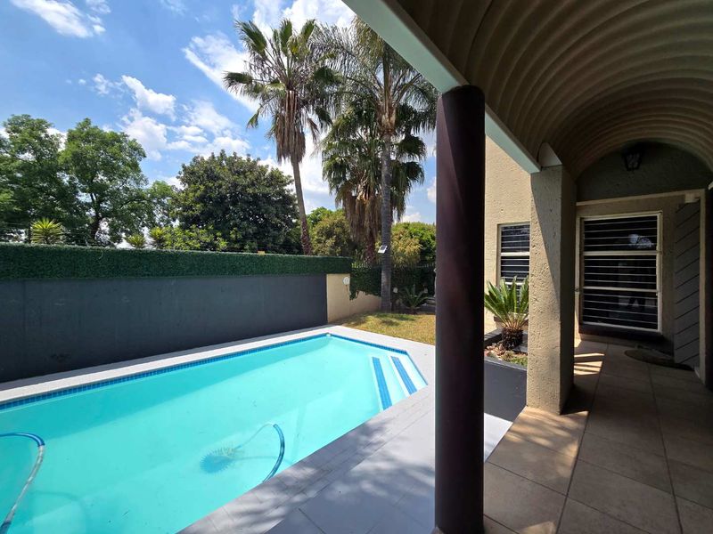 A well-maintained outdoor pool area featuring a tiled patio, covered walkway with corrugated roofing, and a private boundary wall.