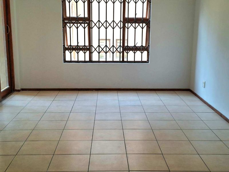 An empty living room featuring neutral floor tiles, a large window with a security gate, and direct access to an outdoor area.