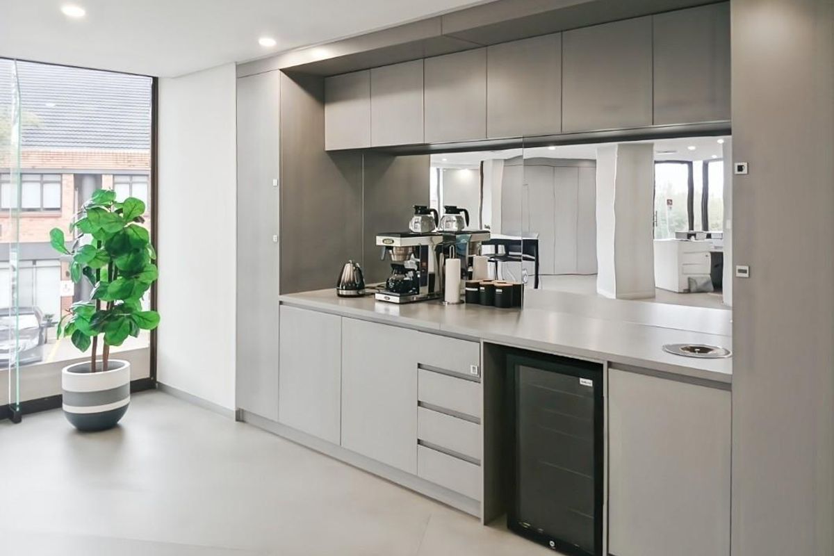 Modern office kitchenette featuring sleek grey cabinetry, a mirrored splashback, integrated beverage fridge, and recessed lighting, creating a clean and professional break area.