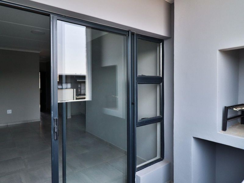 A clean, modern balcony featuring tiled flooring, a built-in braai, and large sliding glass doors providing seamless access to the interior living space.