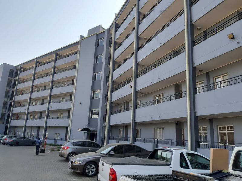 A multi-story apartment building exterior featuring a curved facade with private balconies, metal railings, and an adjacent paved parking area.