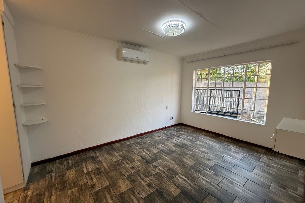A bright, empty bedroom featuring wood-look tiled flooring, a wall-mounted air conditioning unit, built-in shelving, and a window with security bars.