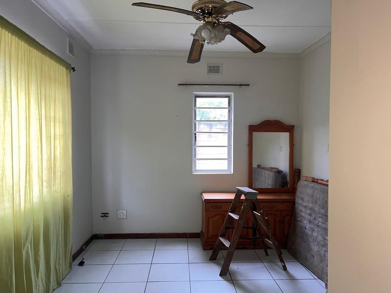A simple bedroom featuring tiled flooring, a ceiling fan, and a window with security bars, offering a blank canvas for renovation.