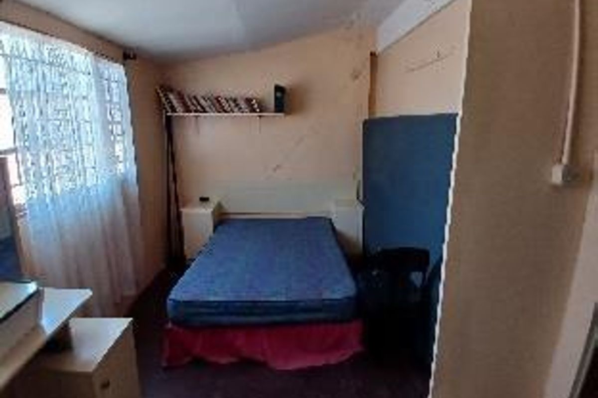 A compact bedroom featuring a built-in headboard, shelving, and large windows with security bars, requiring cosmetic updates to improve the overall finish.