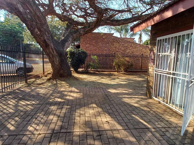 A paved outdoor patio area featuring a large mature tree, brick exterior walls, and a secure metal gate entrance.