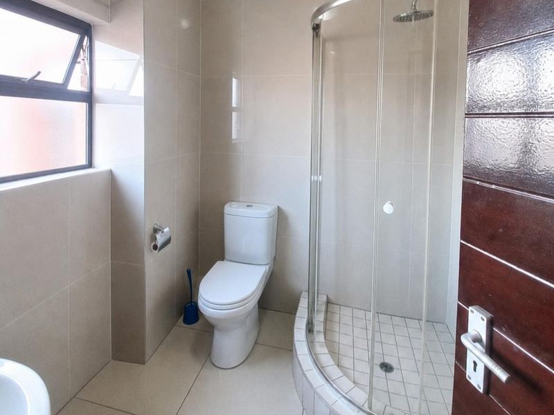 A clean, modern bathroom featuring a curved glass shower enclosure, neutral wall and floor tiling, and a top-hung window for natural light.
