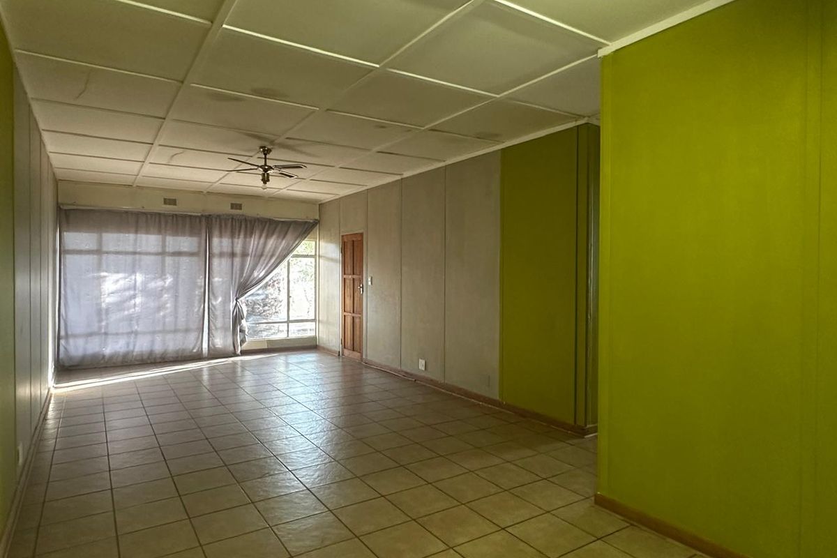 A spacious, empty living area featuring tiled flooring, a drop ceiling with a fan, and large windows allowing natural light.