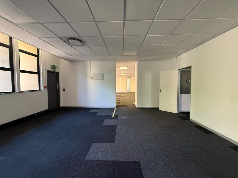 An empty, open-plan office space featuring a suspended ceiling, carpeted flooring, and direct access to adjacent office areas.