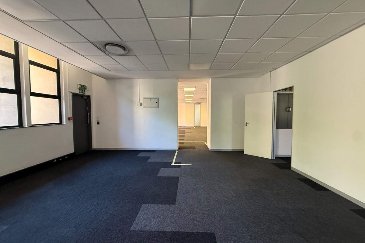 An empty, open-plan office space featuring a suspended ceiling, carpeted flooring, and direct access to adjacent office areas.