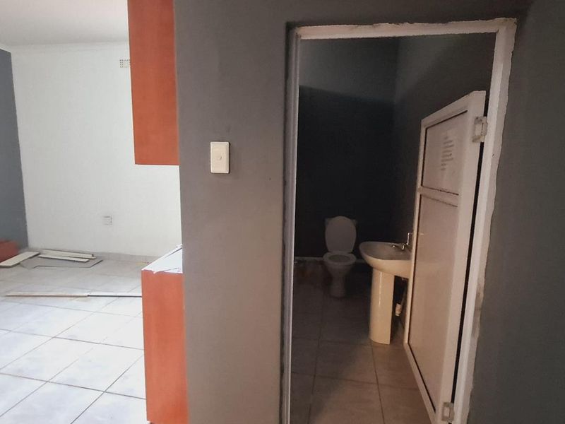A small, unfinished bathroom featuring a pedestal basin and toilet, adjacent to an open-plan living area with tiled flooring.