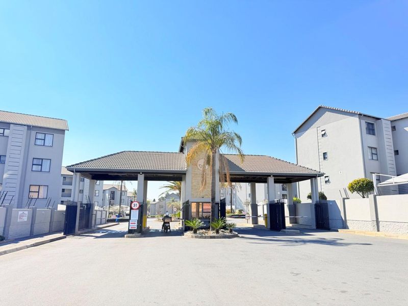 The main entrance and security gatehouse of the apartment complex, featuring a paved driveway, boom gate access, and well-maintained perimeter structures.