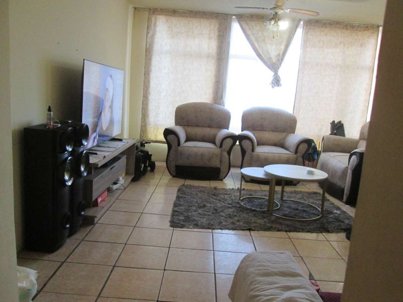 A living room featuring tiled flooring and a ceiling fan, offering a functional space with natural light from large windows.