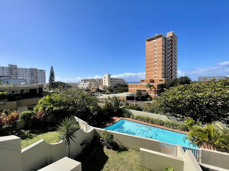 Elevated view of a residential swimming pool area with surrounding garden and boundary walls, set against an urban apartment building backdrop.