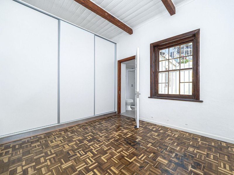 A bright bedroom featuring classic parquet flooring, exposed timber ceiling beams, a large built-in wardrobe, and direct access to an en-suite bathroom.
