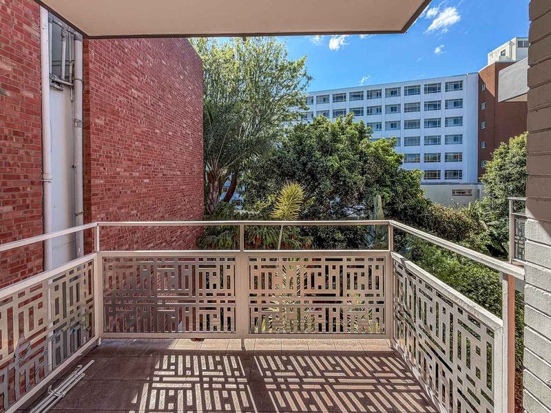 A private balcony featuring a decorative metal balustrade, tiled flooring, and views of the surrounding urban greenery and neighboring buildings.