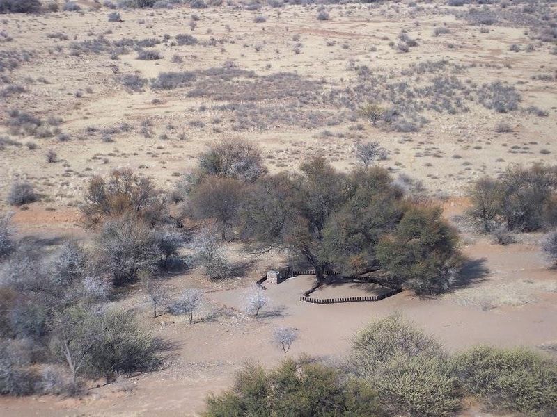 An aerial view of a rural farm landscape featuring a small stone masonry structure and timber pole enclosure nestled among native trees.