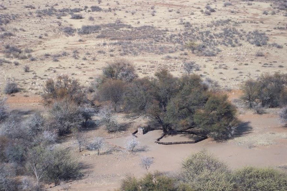 An aerial view of a rural farm landscape featuring a small stone masonry structure and timber pole enclosure nestled among native trees.