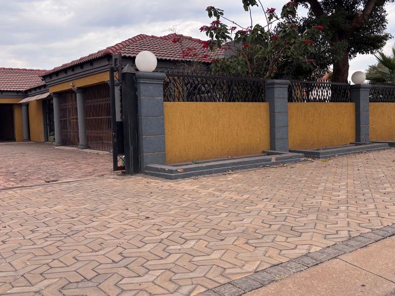 Front exterior view of the property featuring a paved driveway, secure boundary wall with decorative pillars, and a tiled roof structure.