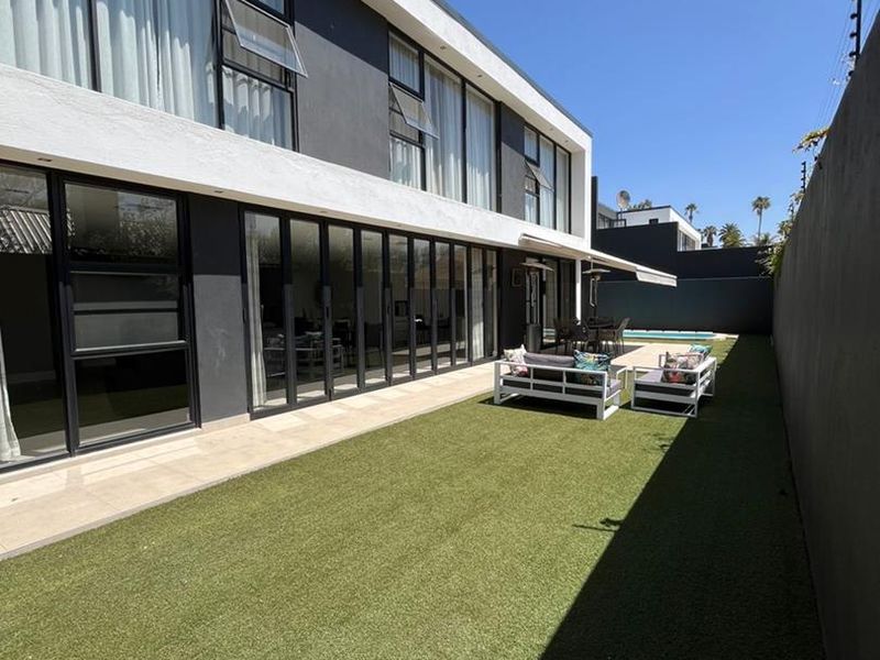Modern rear exterior featuring a manicured artificial lawn, large bi-fold doors leading to the interior, a retractable awning, and a private swimming pool.