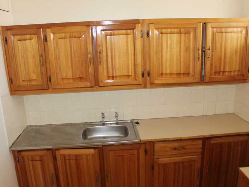 A compact kitchen area featuring traditional wooden cabinetry, a stainless steel sink, and a tiled backsplash, offering a functional but dated layout.