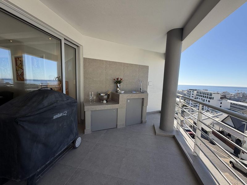 A spacious covered balcony featuring a built-in kitchenette with a sink, tiled flooring, and a metal balustrade, offering expansive coastal views.