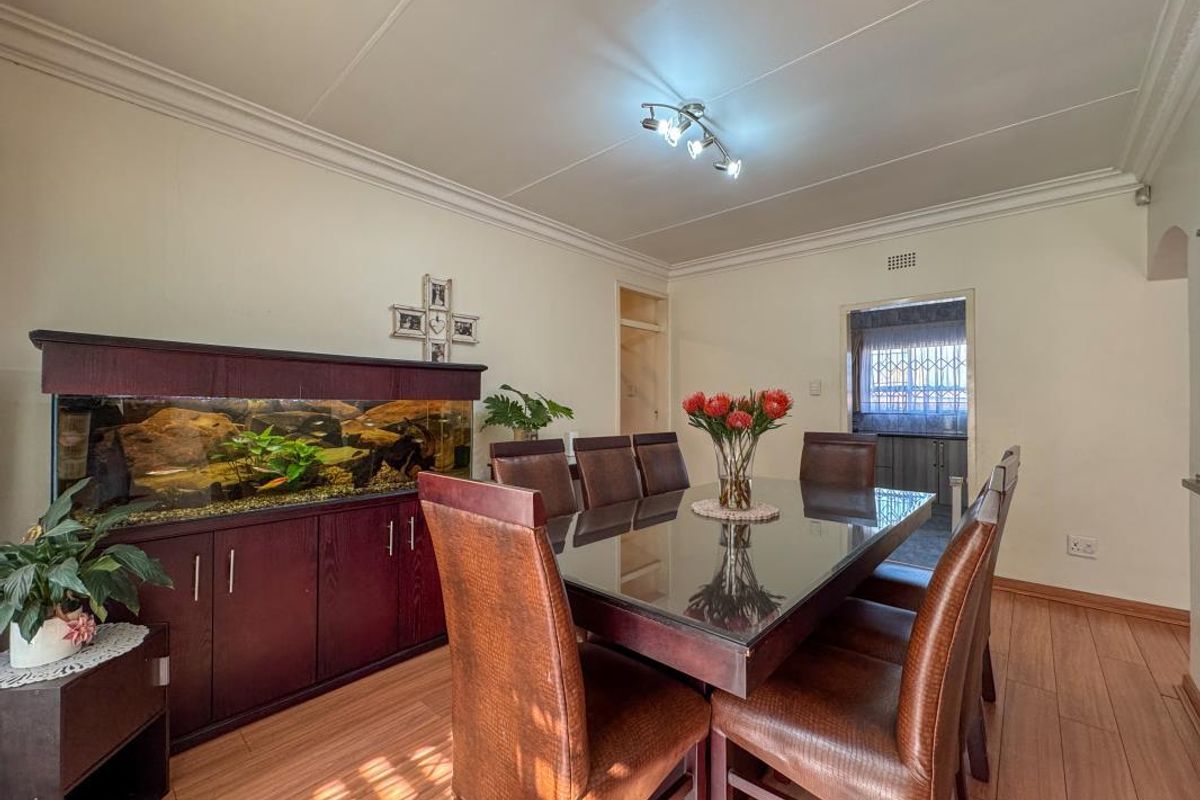 A dining room featuring laminate flooring, ceiling cornice, and a central light fixture, with an adjacent view into a secondary kitchen area.