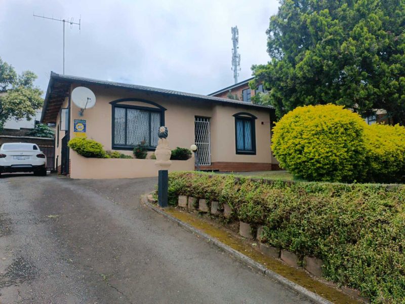 Front elevation of a single-story house featuring an asphalt driveway, landscaped garden, and security gate entrance.