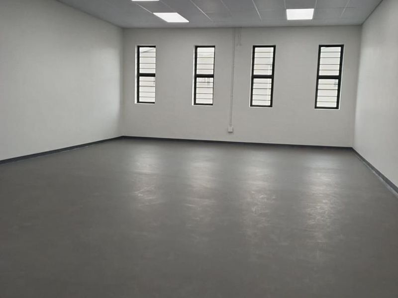 A clean, vacant industrial office space featuring neutral walls, durable flooring, recessed ceiling lighting, and windows fitted with security bars.