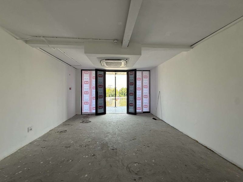An empty retail space featuring a ceiling-mounted air conditioning unit, double glass doors, and concrete flooring in need of refurbishment.