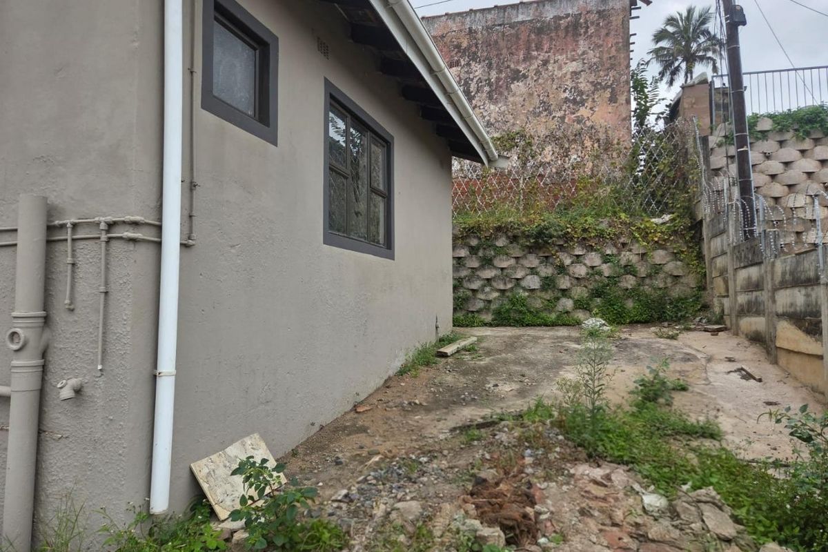 Rear exterior view of the property showing a concrete area, retaining wall, and security fencing, requiring maintenance and landscaping.