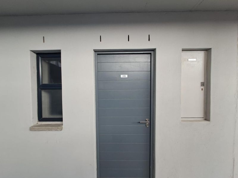Apartment entrance featuring a grey door, adjacent window, and utility access panel in a neutral wall finish.
