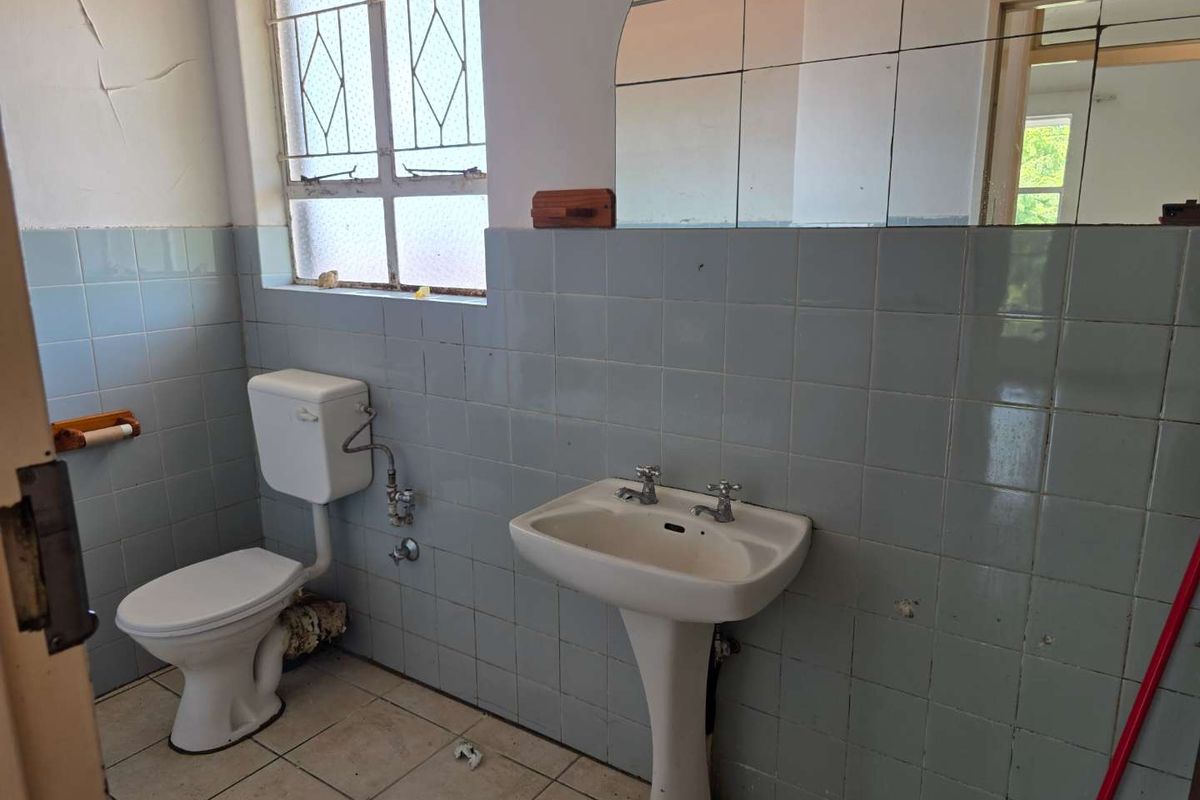 A dated bathroom featuring a pedestal basin, standard toilet, and wall-mounted mirror, requiring significant renovation and cosmetic updates to modernize the space.
