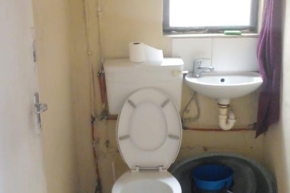 A compact bathroom featuring a standard toilet, wall-mounted basin, and tiled flooring, requiring cosmetic updates and finishing of exposed plumbing.