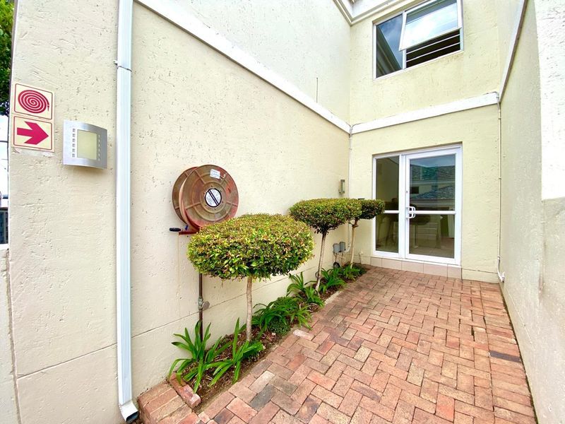 A paved rear courtyard area featuring a fire hose reel, landscaped shrubs, and access to the apartment via a glass sliding door.